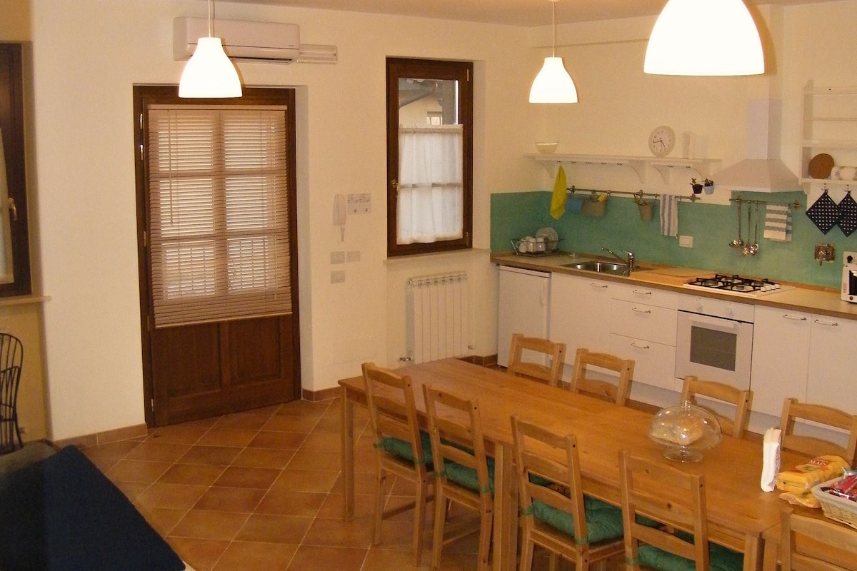 A dining area with a wooden table and chairs, a sofa with cushions, and a kitchen in the background.