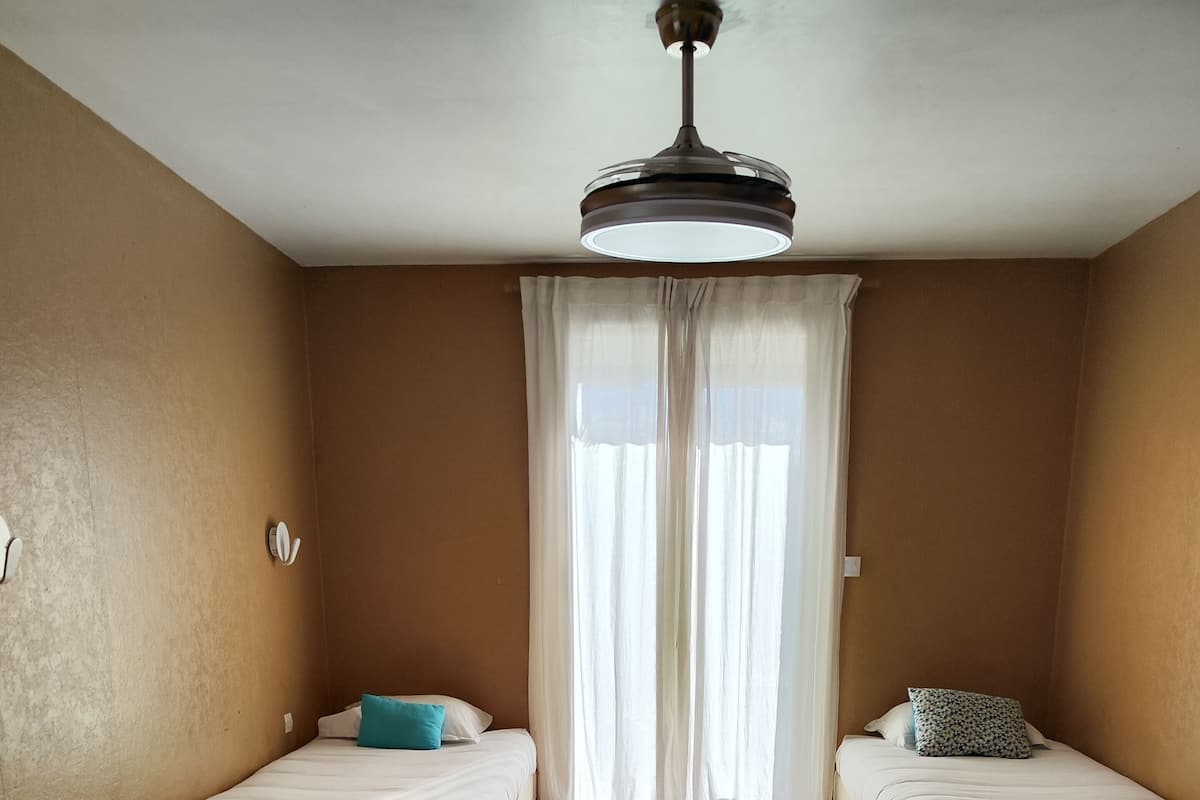 A room with two single beds, a ceiling fan, and a window with sheer curtains.
