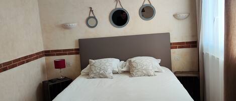 A neatly made bed with white linens and decorative pillows, a bedside table with a lamp, and wall-mounted mirrors and sconces.