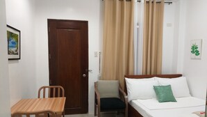 A small, neatly arranged room with a wooden door, a bed with white bedding and a green pillow, a small table with chairs, and a window with beige curtains. - Lighthouse Apartment and Guest Inn (Puerto Princesa)