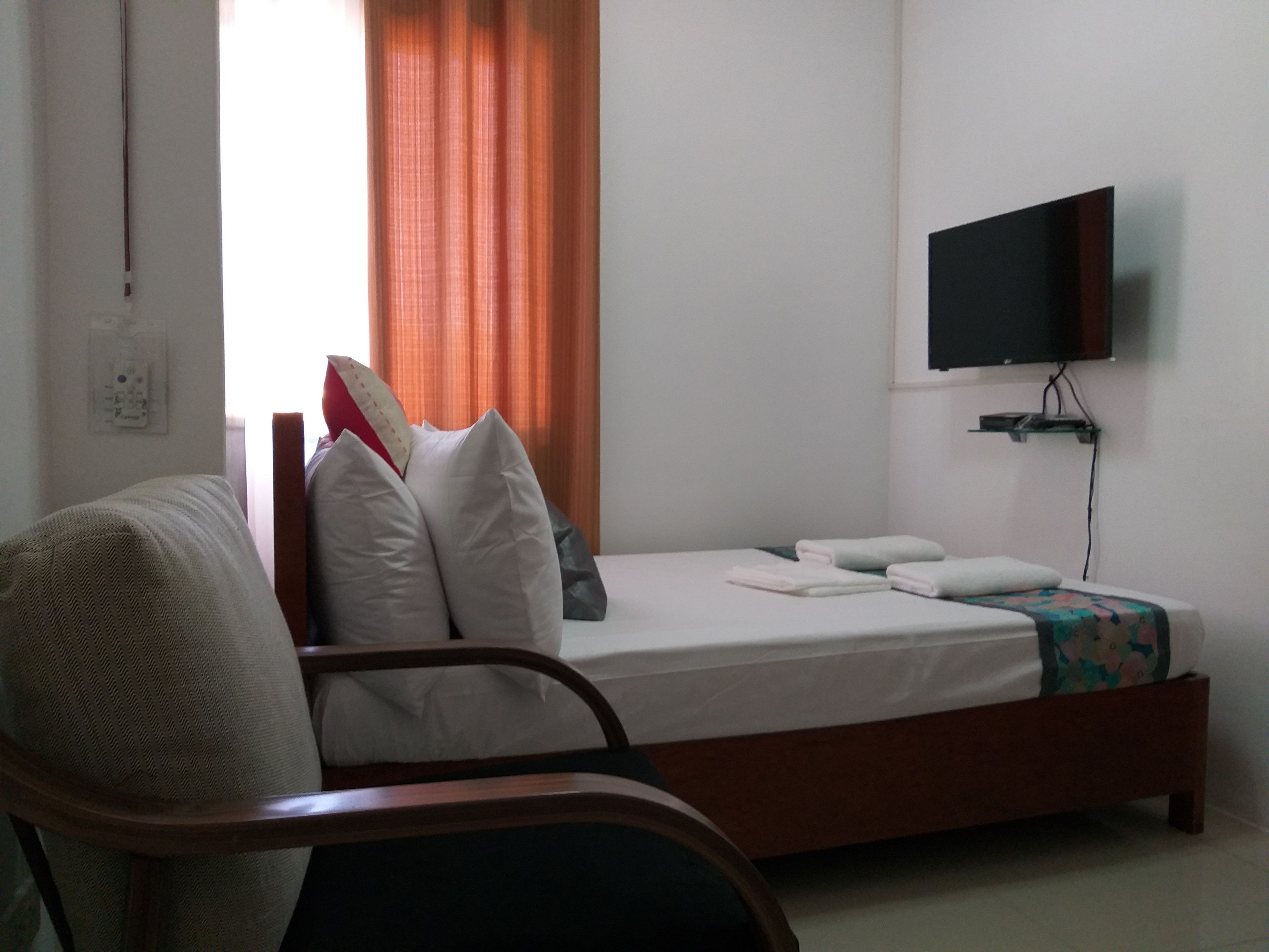 Un dormitorio con cama, una silla, televisor en la pared y ventana con cortinas.