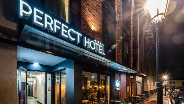 Das äußere Erscheinungsbild des Perfect Hotels mit einem offenen Eingangsbereich, einer Außenbestuhlung und einem Parkplatz.