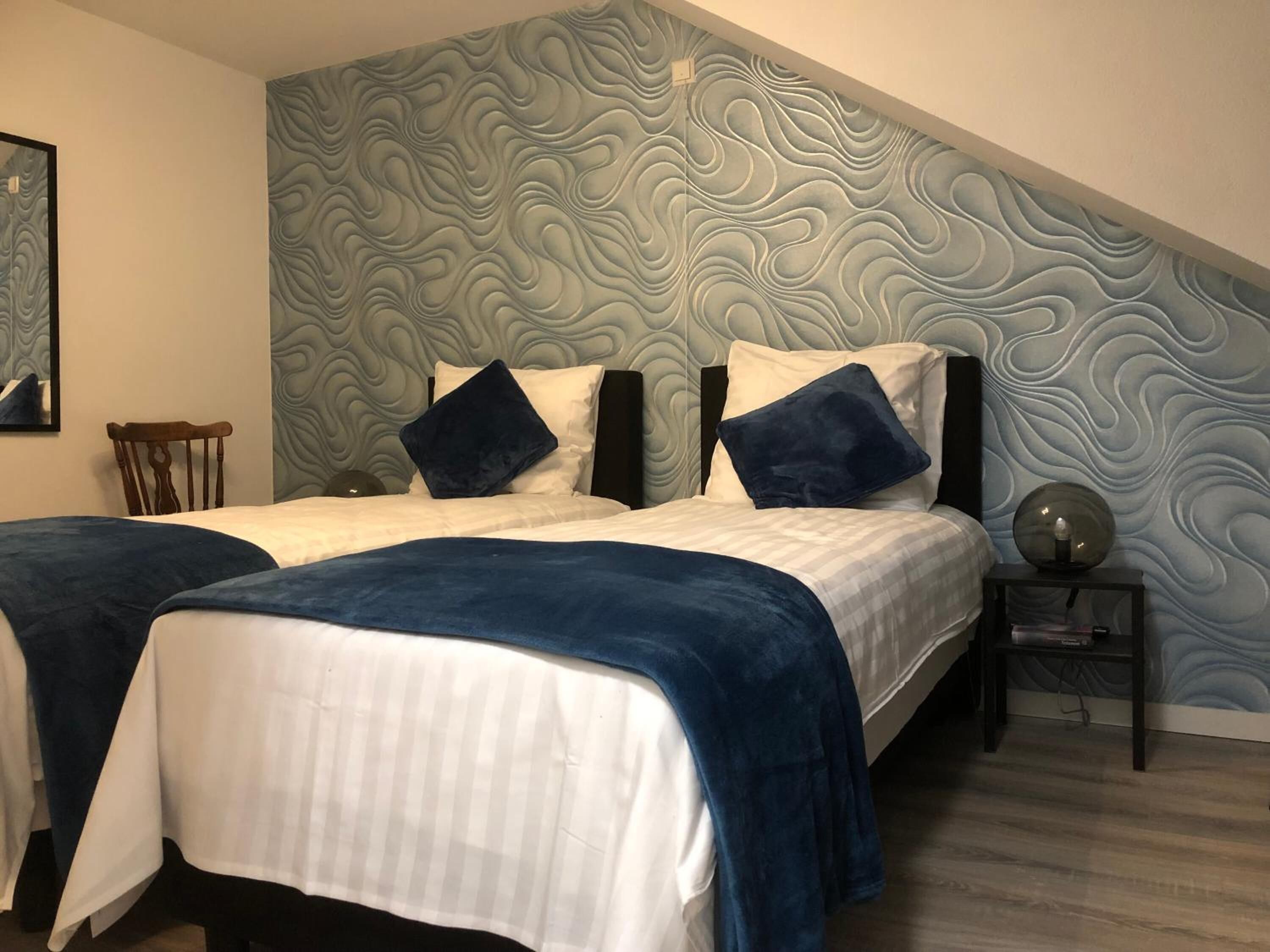 Un dormitorio con dos camas, una silla de madera, una mesita de noche con una lámpara y una pared con diseño ondulado.