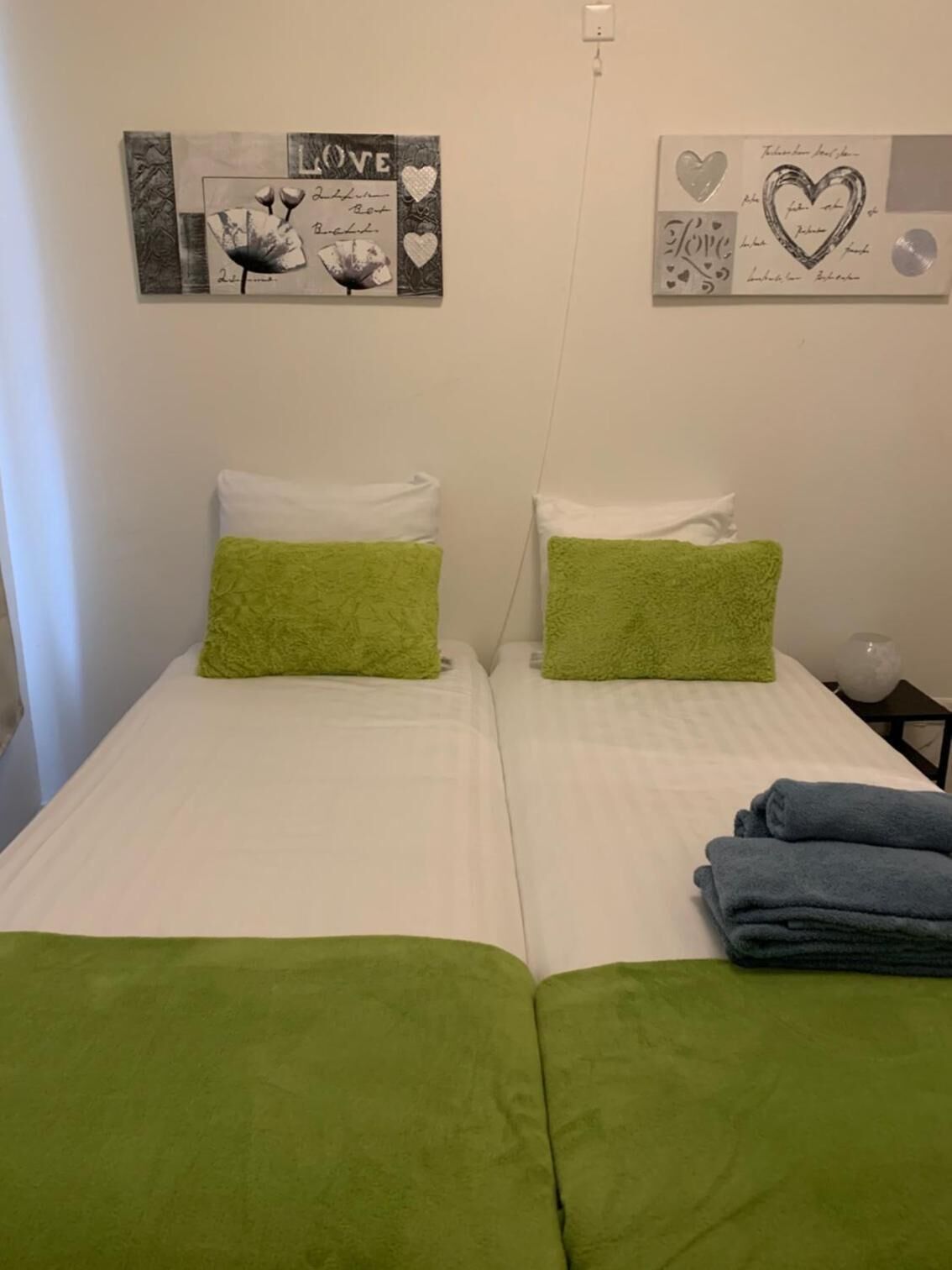 Een kamer met twee eenpersoonsbedden, elk met een groene kussen en een witte kussen. Twee kunstwerken hangen aan de muur, waarvan één de tekst 'LOVE' bevat en de ander harten en de tekst 'Love'.