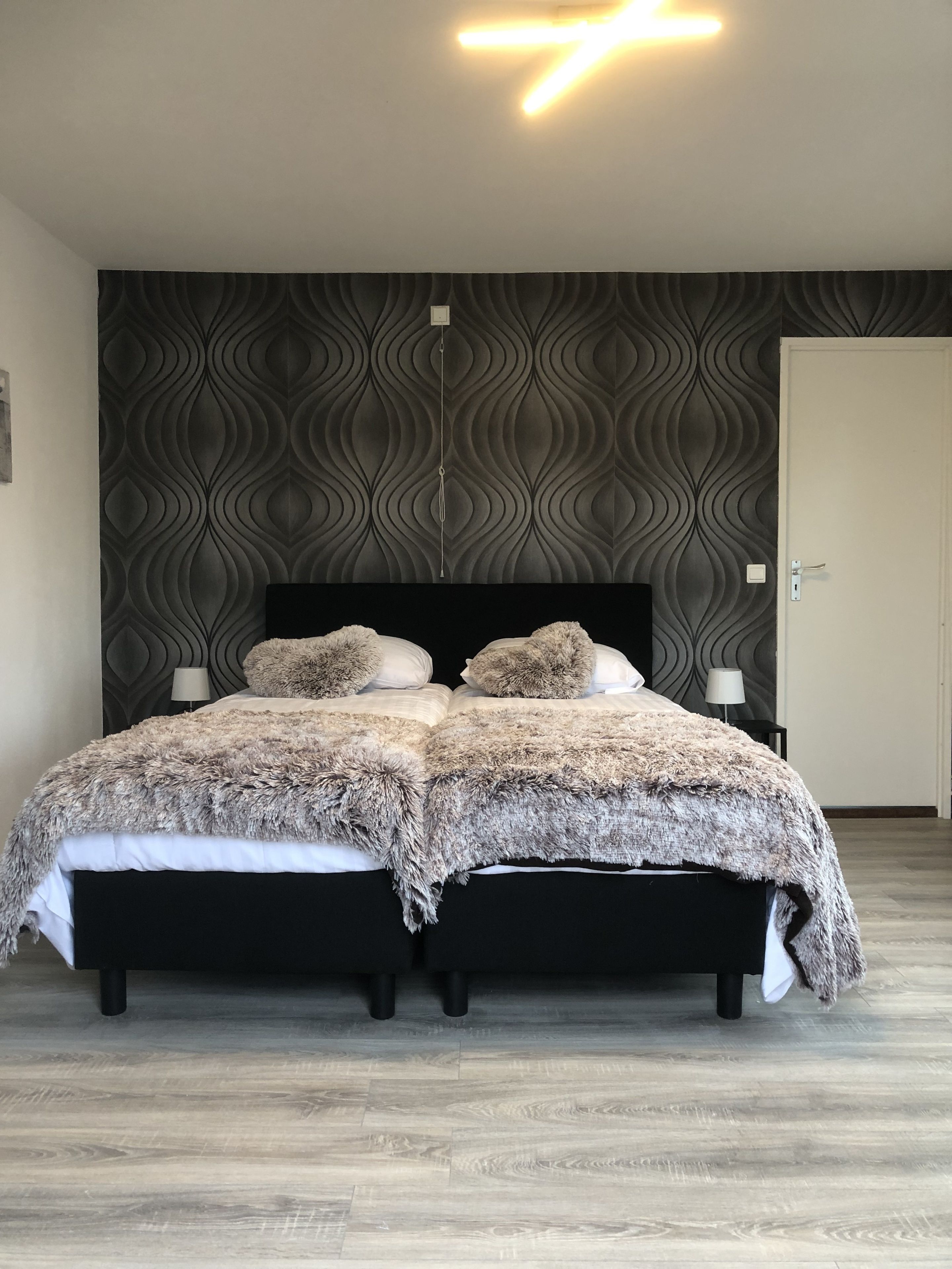 Un lit double avec une couverture moelleuse et deux oreillers, une table de chevet avec une lampe, et un mur orné d’un motif ondulé.