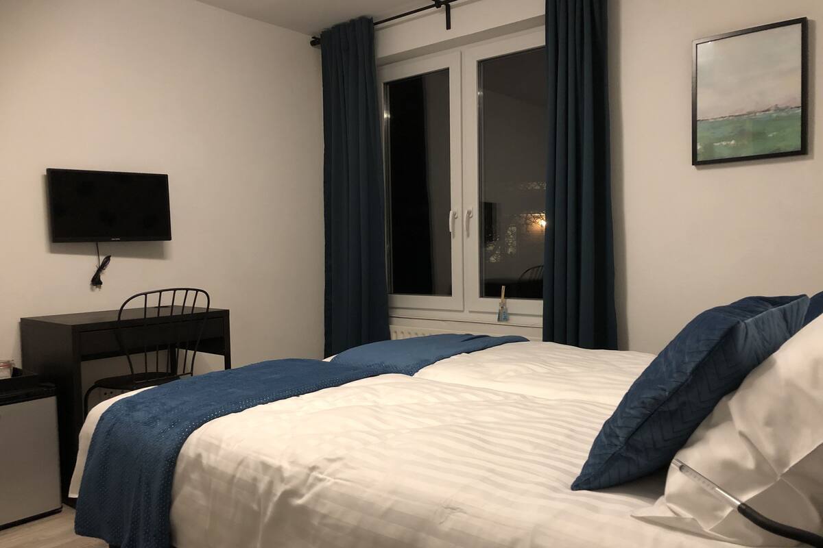 A bedroom with a bed, a desk, a chair, a TV, and a window with curtains.