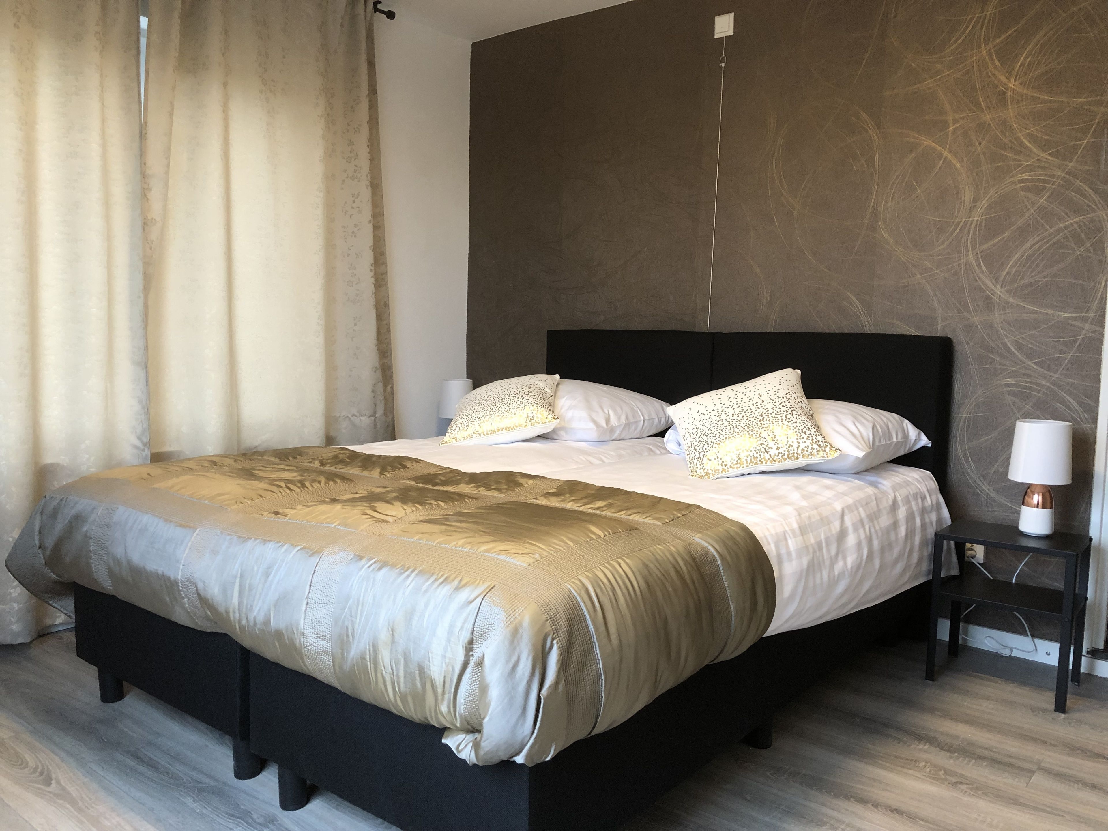 A neatly made bed with a dark headboard, a bedside table with a lamp, and a curtain partially drawn to the side.