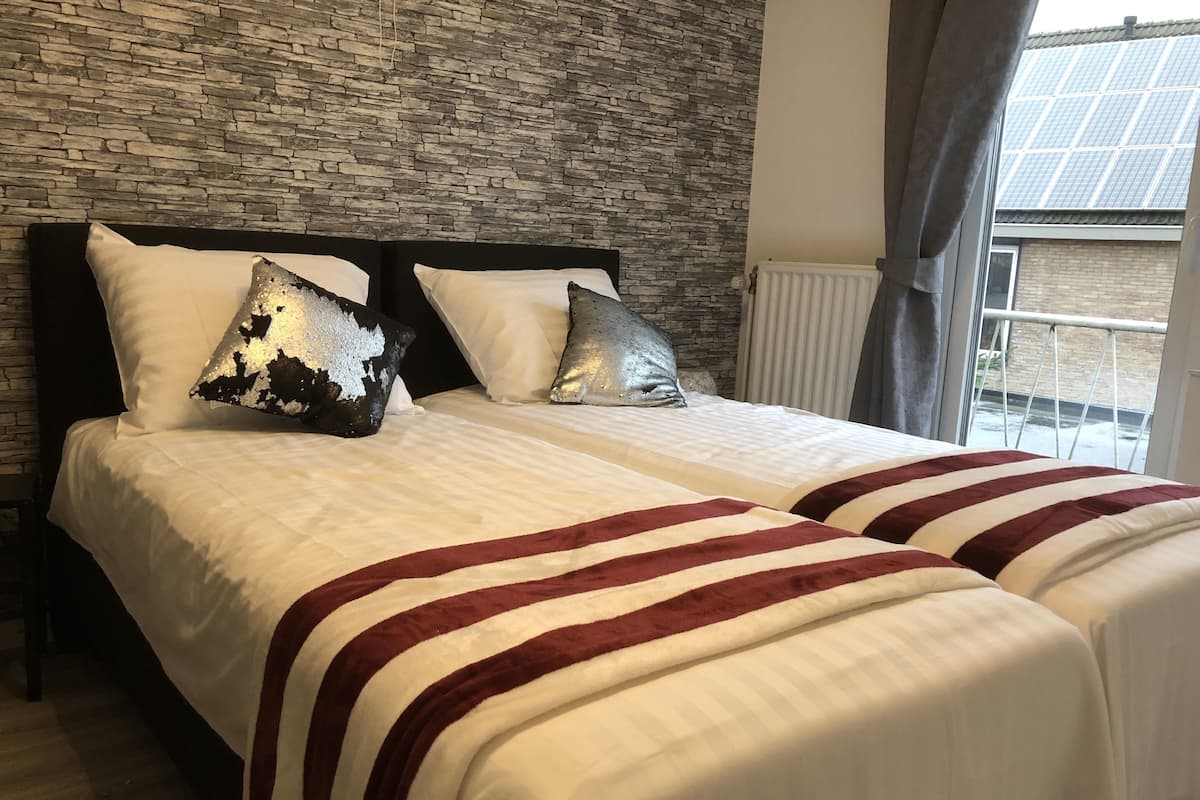 A double bed with white linens and red stripes, two pillows, a stone wall, and a window with curtains.