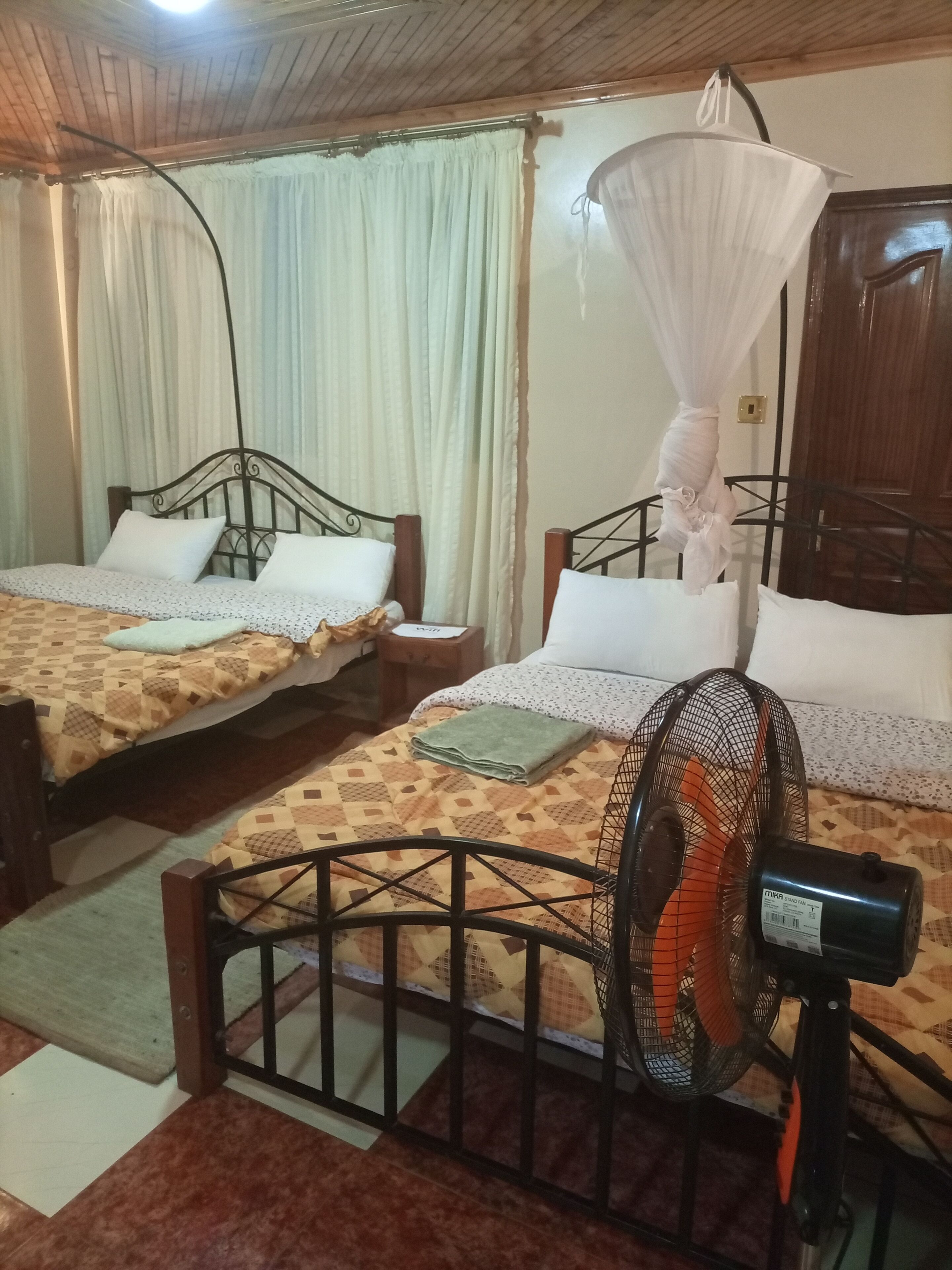 A room with two beds, each with a mosquito net, a standing fan, and a wooden wardrobe.