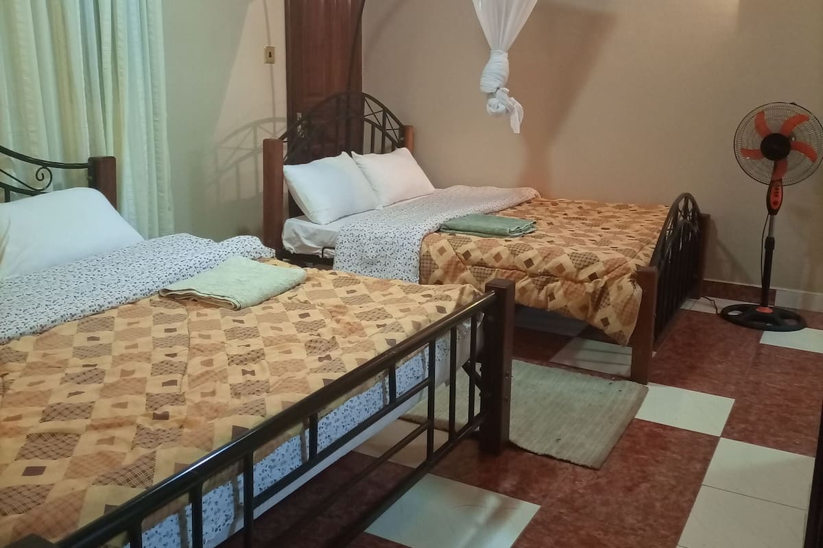 Une chambre avec deux lits simples, un ventilateur sur pied, une moustiquaire et un sol carrelé.