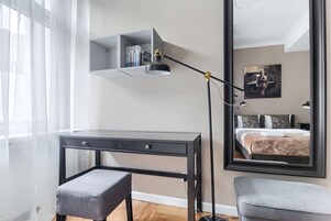 A modern bedroom with a desk, a bench, a bookshelf, and a large mirror reflecting a bed and artwork.