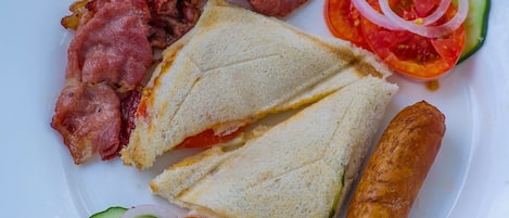 A plate with a sandwich, bacon, sausage, and sliced vegetables.