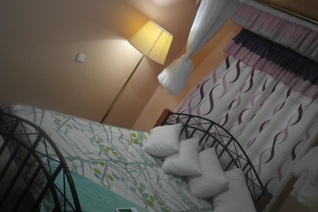 A bedroom with a bed, a bedside lamp, a window with curtains, and a wall-mounted thermostat.
