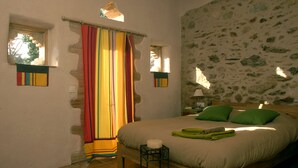 Un dormitorio con una cama, almohadas verdes, una mesita de noche, una lámpara, un ventanal y una cortina colorida.