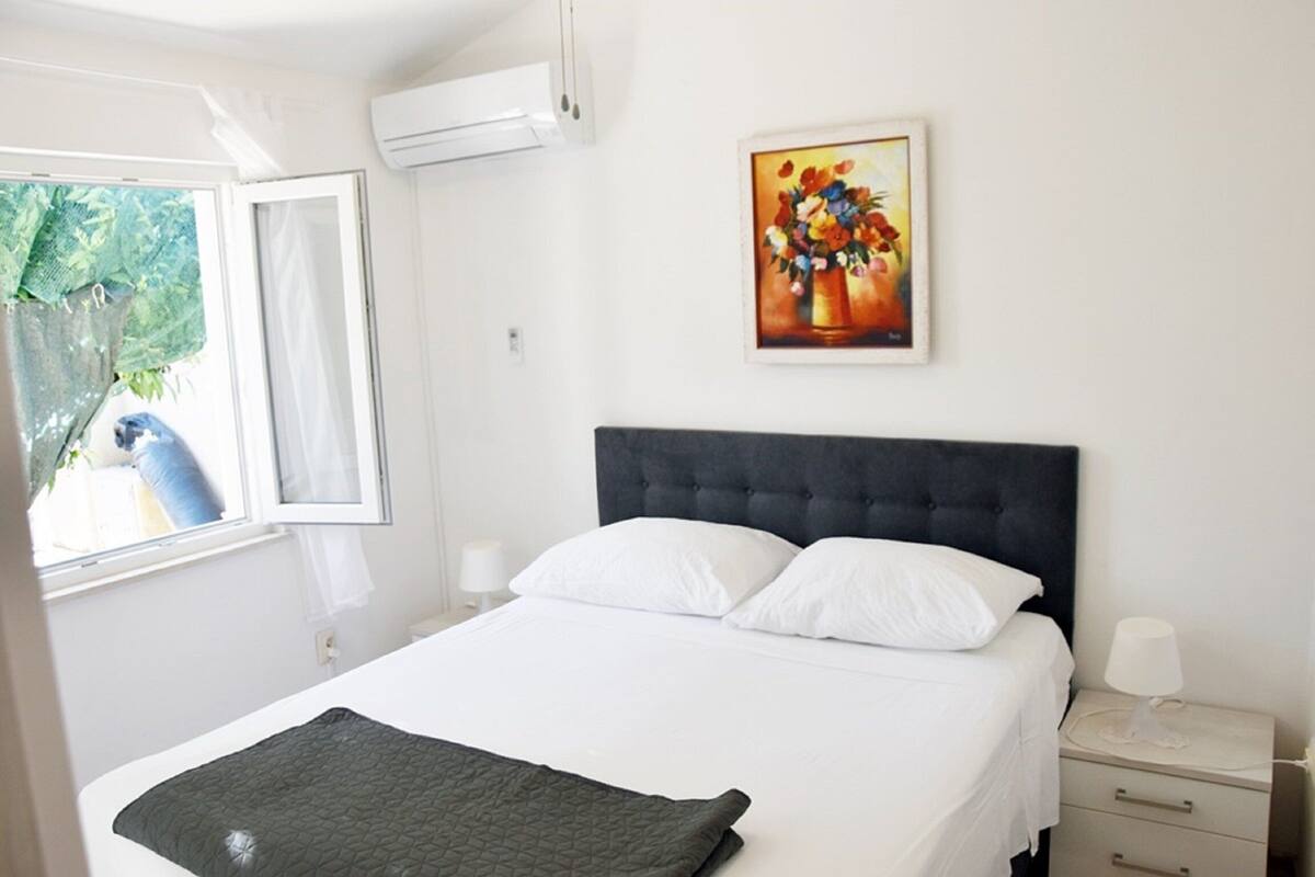 A neatly made bed with white bedding and a dark headboard, a bedside table with a lamp, and a painting of flowers on the wall.