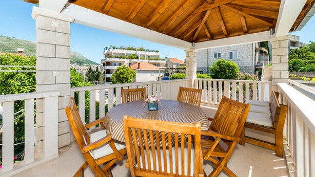 Un patio techado al aire libre con una mesa redonda y sillas de madera, con vistas a una colina con edificios.