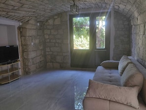 Habitación con paredes de piedra, un televisor sobre un soporte, un sofá con cojines y una puerta que ofrece vistas a la vegetación exterior.