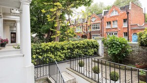Un quartier résidentiel comprenant un immeuble blanc, une maison en briques rouges et un jardin bien entretenu, agrémenté d’une clôture et de plantes en pot.