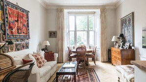Un salon traditionnel avec un canapé blanc, une table basse en verre, une table à manger avec des chaises, un tapis à motifs et une grande fenêtre avec des rideaux.