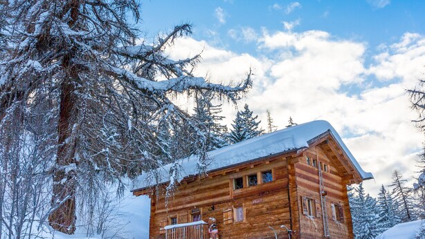 Une cabane en bois dont le toit est recouvert de neige, entourée d’arbres chargés de neige et sous un ciel dégagé.