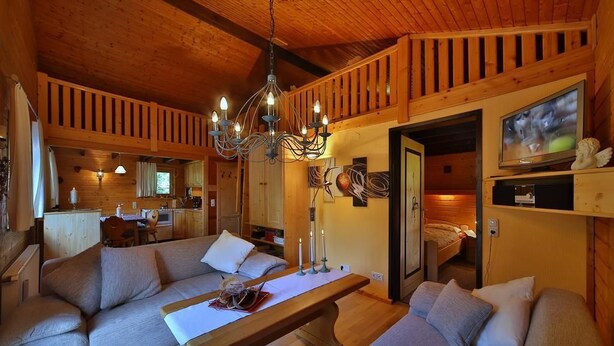 A cozy cabin interior with a wooden ceiling, a dining area with a table and benches, a kitchen in the background, and a television on a shelf. - Ferienhaus Mitterdorf (Philippsreut)