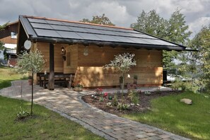 A wooden cabin with a sloped roof, a small porch, and a garden with various plants and flowers. - Ferienhaus Mitterdorf (Philippsreut)