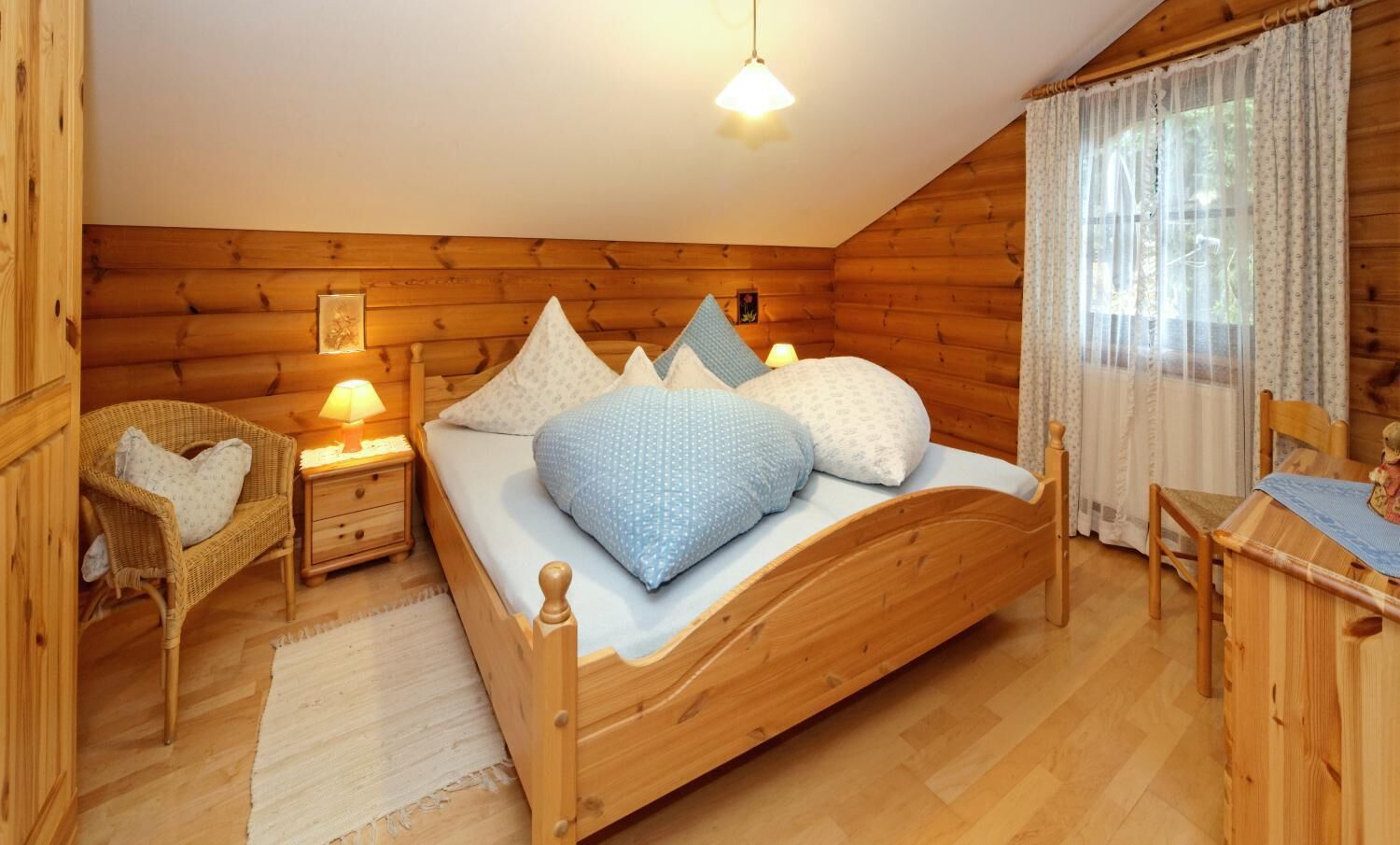 A cozy wooden cabin bedroom with a wooden bed, a nightstand with a lamp, a wicker chair, and a small table with a chair.