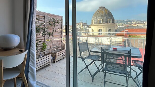 A modern room with a large sliding glass door leading to an outdoor terrace. The terrace features a table with chairs and a view of a historic building with a dome. - Naples from a terrace - 6 (Napoli)