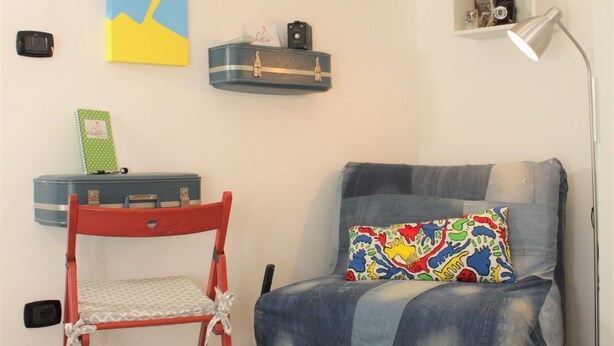 Una habitación con una silla roja, un sofá azul con un cojín colorido, un televisor de pantalla plana, un aire acondicionado, una estantería con libros y un escritorio con una lámpara.
