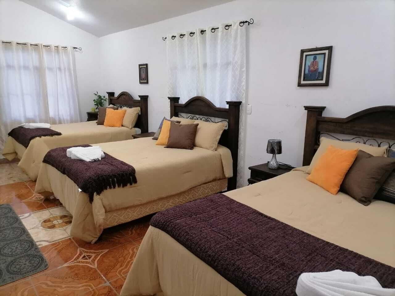 Une chambre d’hôtel avec trois lits, chacun doté d’une tête de lit en bois, de draps beiges et d’un plaid violet. On y trouve une petite table de chevet avec une lampe, un tableau encadré au mur et une fenêtre avec des rideaux blancs.