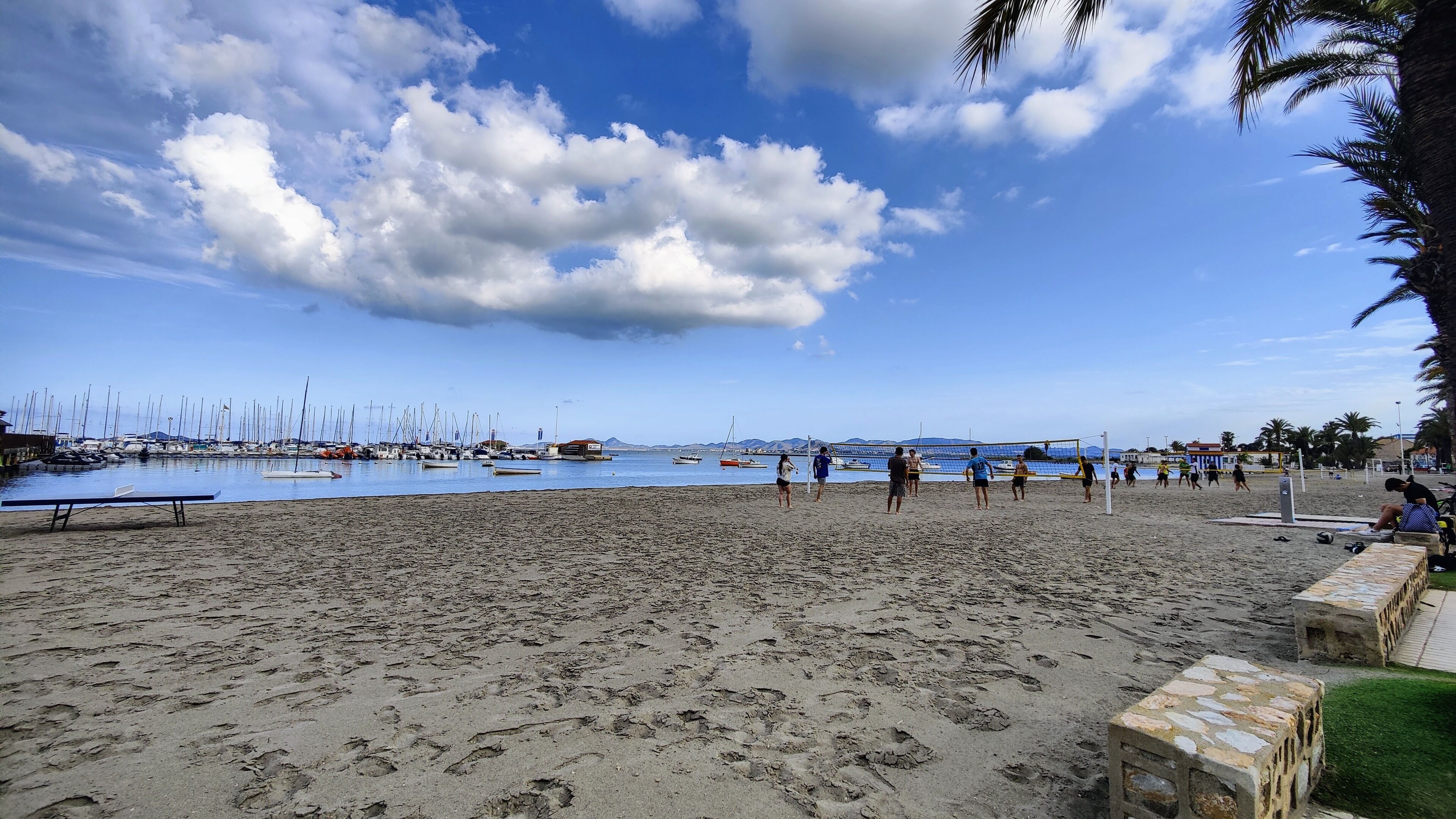 Una playa con un partido de voleibol, barcos al fondo y un cielo despejado.