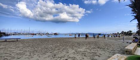 Una playa con un partido de voleibol, barcos al fondo y un cielo despejado.