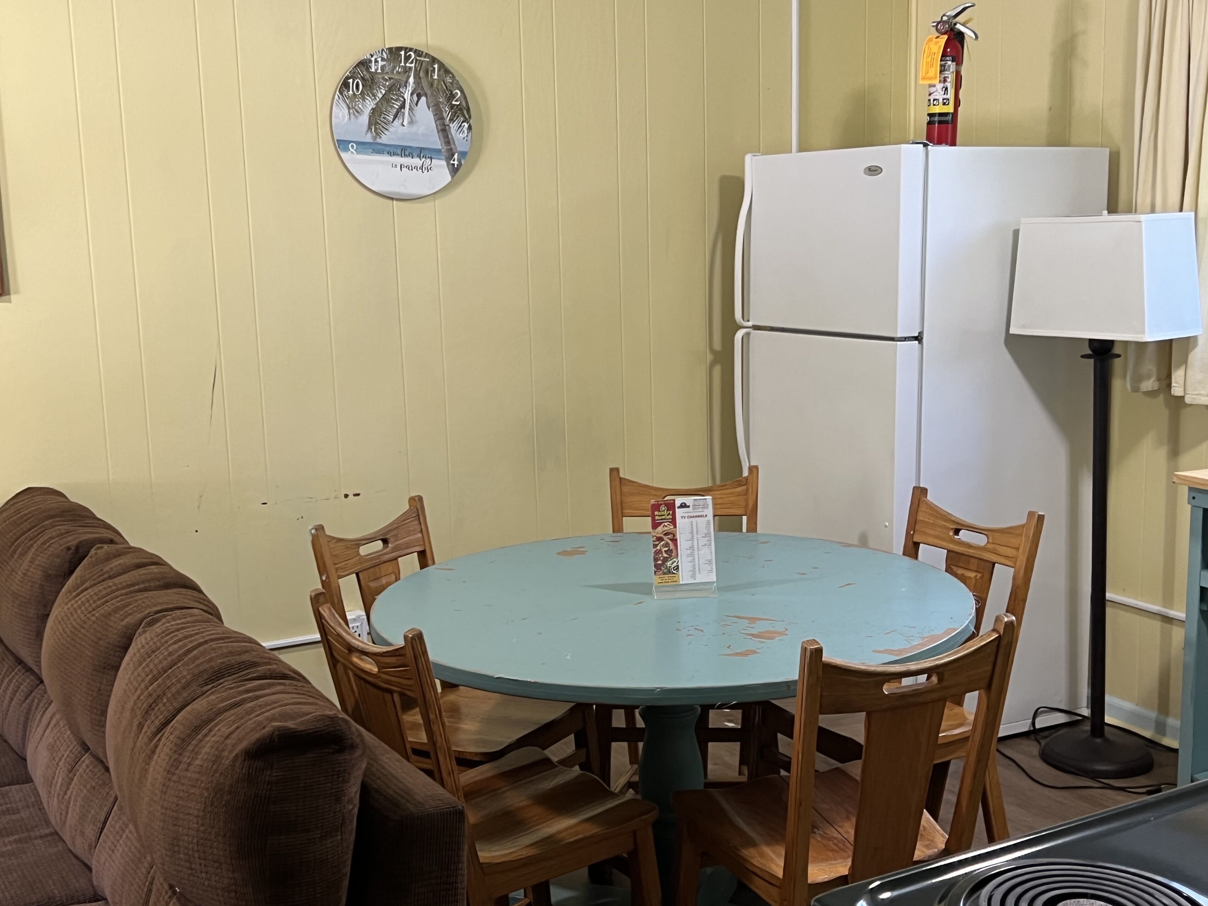 Ein kleiner Essbereich mit einem runden Tisch, Holzstühlen, einer braunen Couch, einem weißen Kühlschrank und einer Wanduhr.