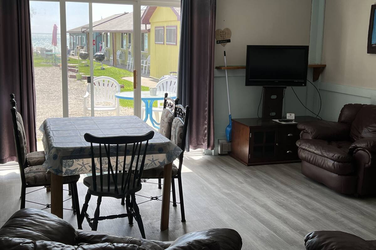 A living room with a dining table, chairs, a sofa, a television, and a view of a beach and buildings outside.