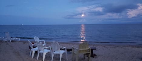 Une plage avec des chaises en plastique blanches et une table en bois, une mer calme et le reflet visible de la lune sur l’eau.