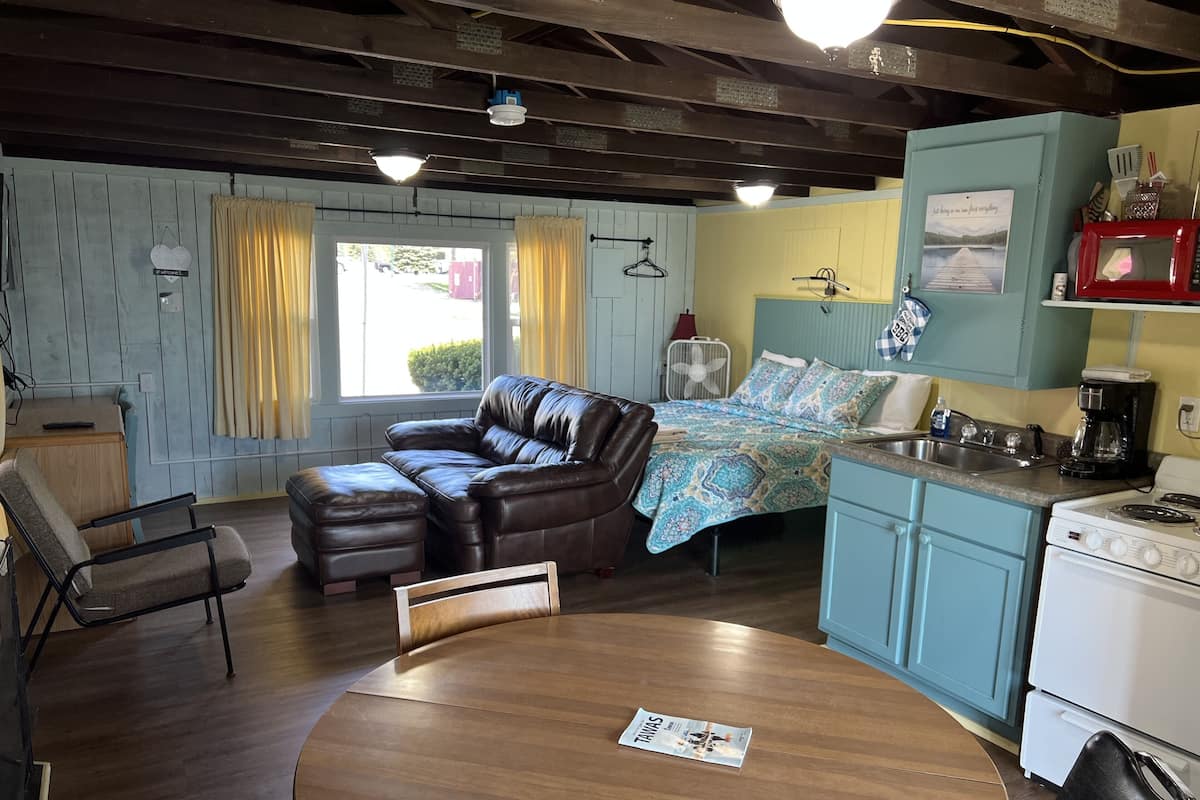 A cozy room with a leather sofa, a dining table, a kitchenette, and a bed with a patterned blanket.