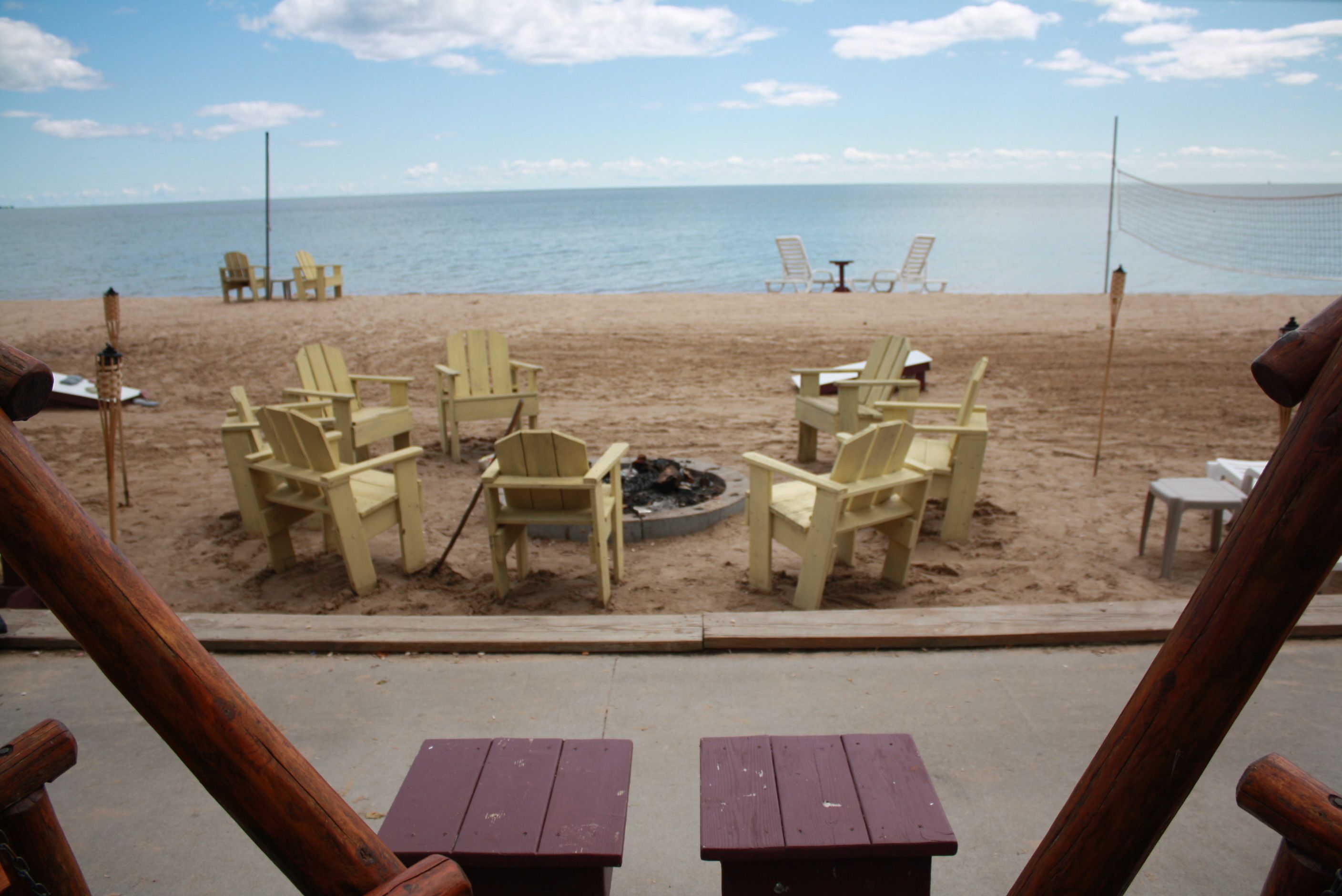 Ein Strand mit Holzhöckern und Tischen zum Sitzen, einem Volleyballnetz und Blick auf das Meer.