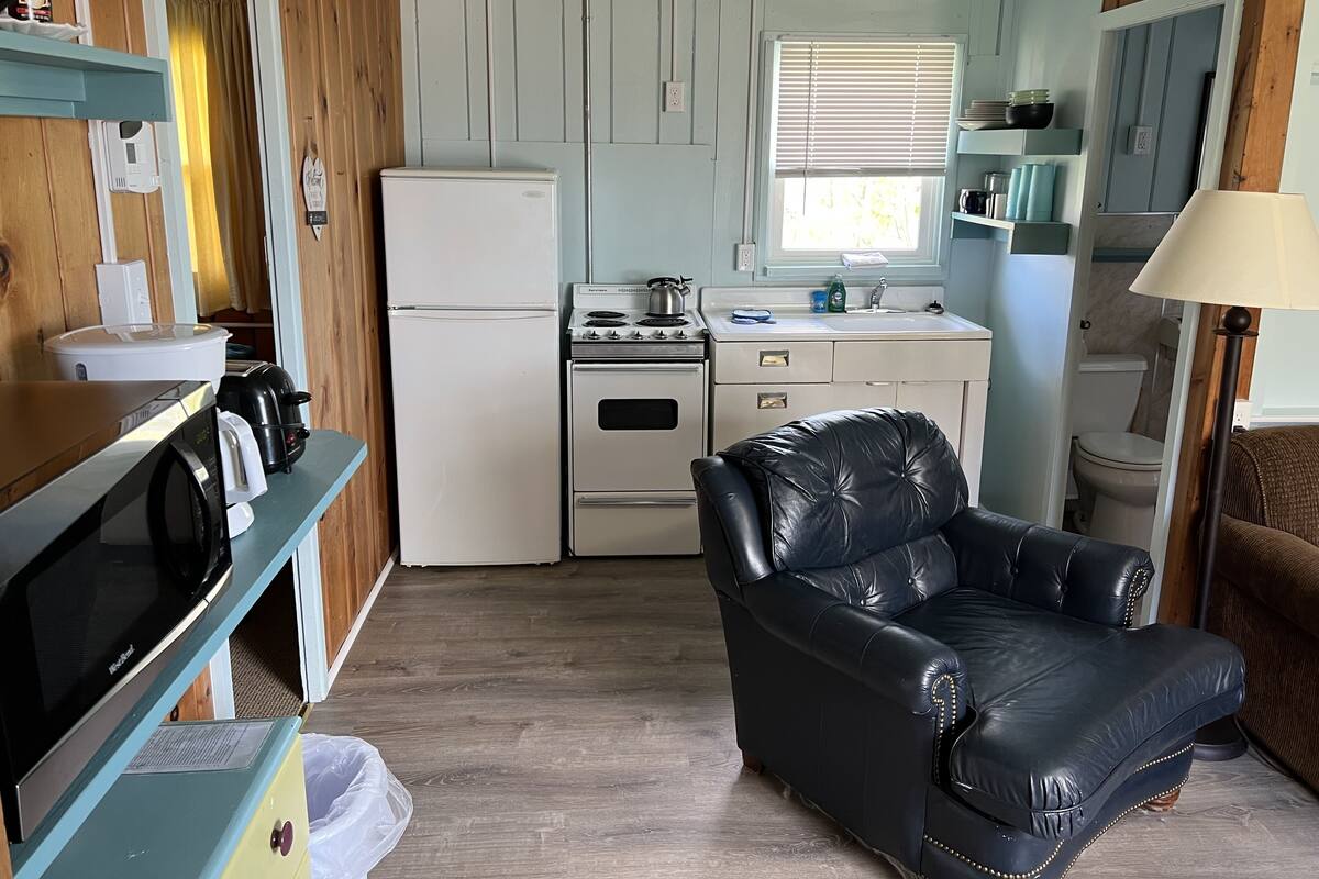 A compact kitchen with a refrigerator, stove, and sink. A black leather chair is in the foreground, and a microwave is on a shelf.