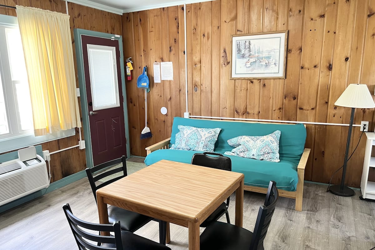 A cozy room with a wooden paneled wall, a teal sofa with patterned cushions, a wooden table with two chairs, and a door with a window and curtains.