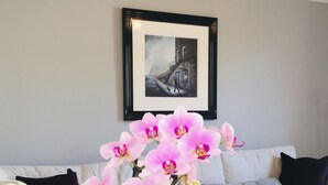 Ein Wohnzimmer mit einem beigen Sofa, einem gerahmten Bild an der Wand und pinkfarbenen Orchideen in einer Vase.