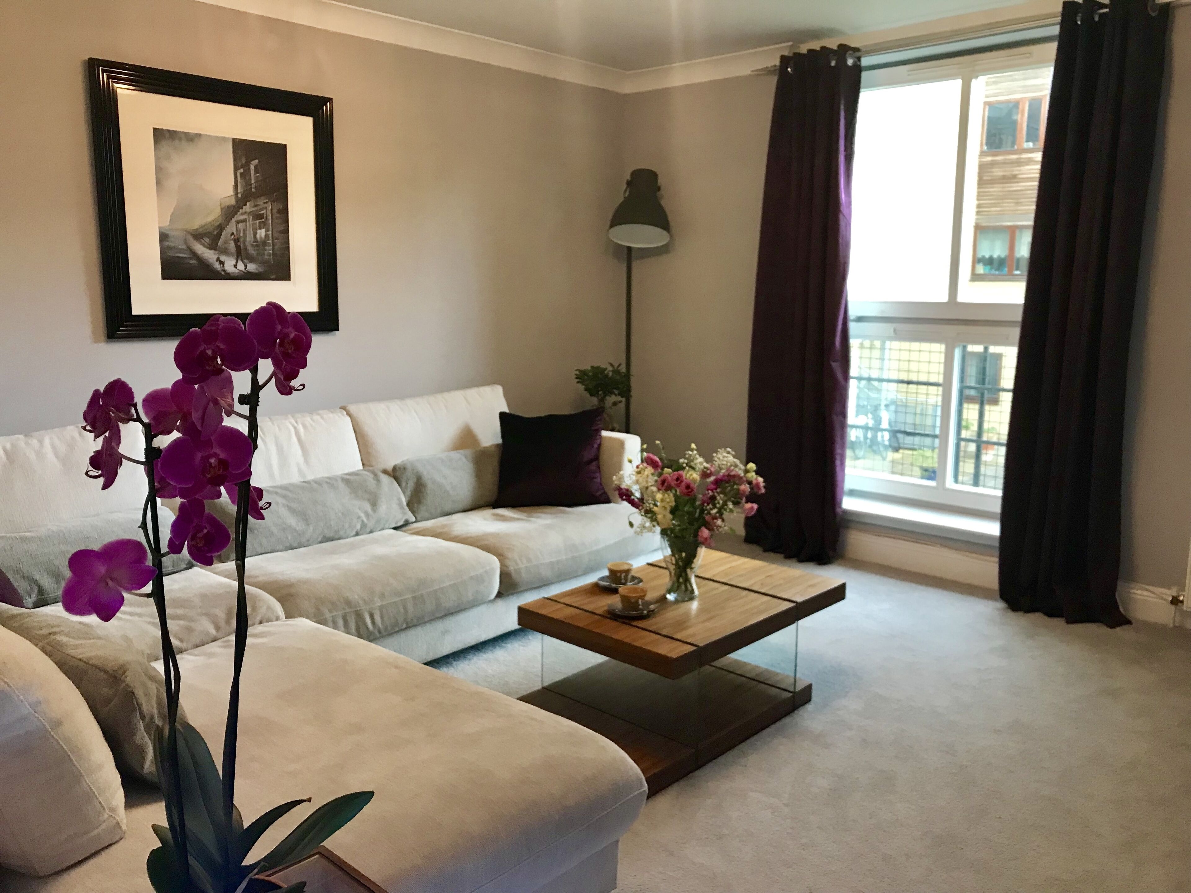 A living room with a sofa, coffee table, orchid plant, framed picture, and a window with curtains.