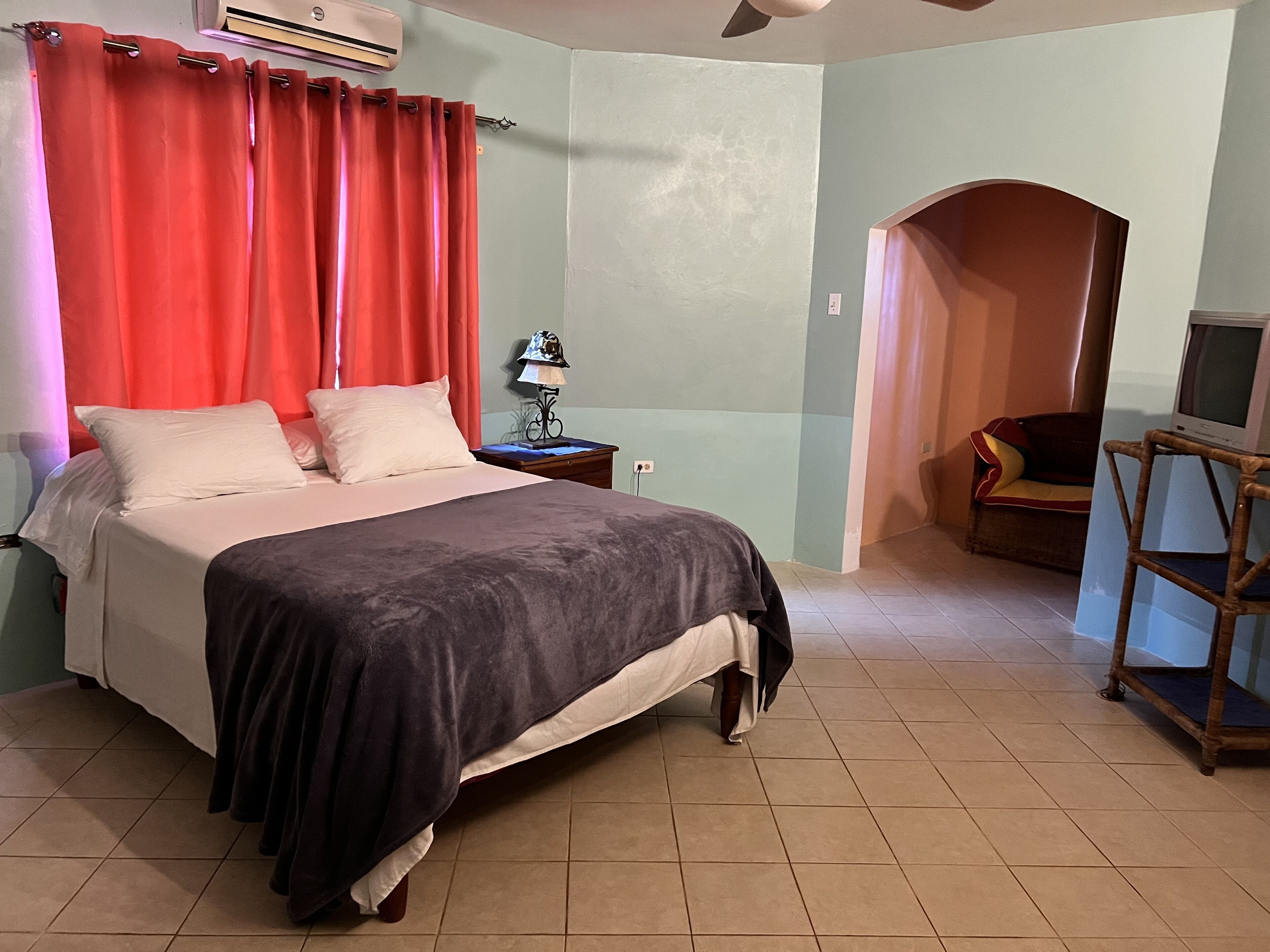 A bedroom with a bed, red curtains, a nightstand with a lamp, a TV on a wooden stand, and a small seating area with a couch.
