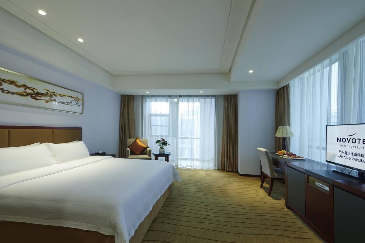 Ein Hotelzimmer mit einem großen Bett, einem Schreibtisch mit Fernseher, einem Sessel, einem kleinen Tisch mit Pflanze und einem Fenster mit Vorhängen.