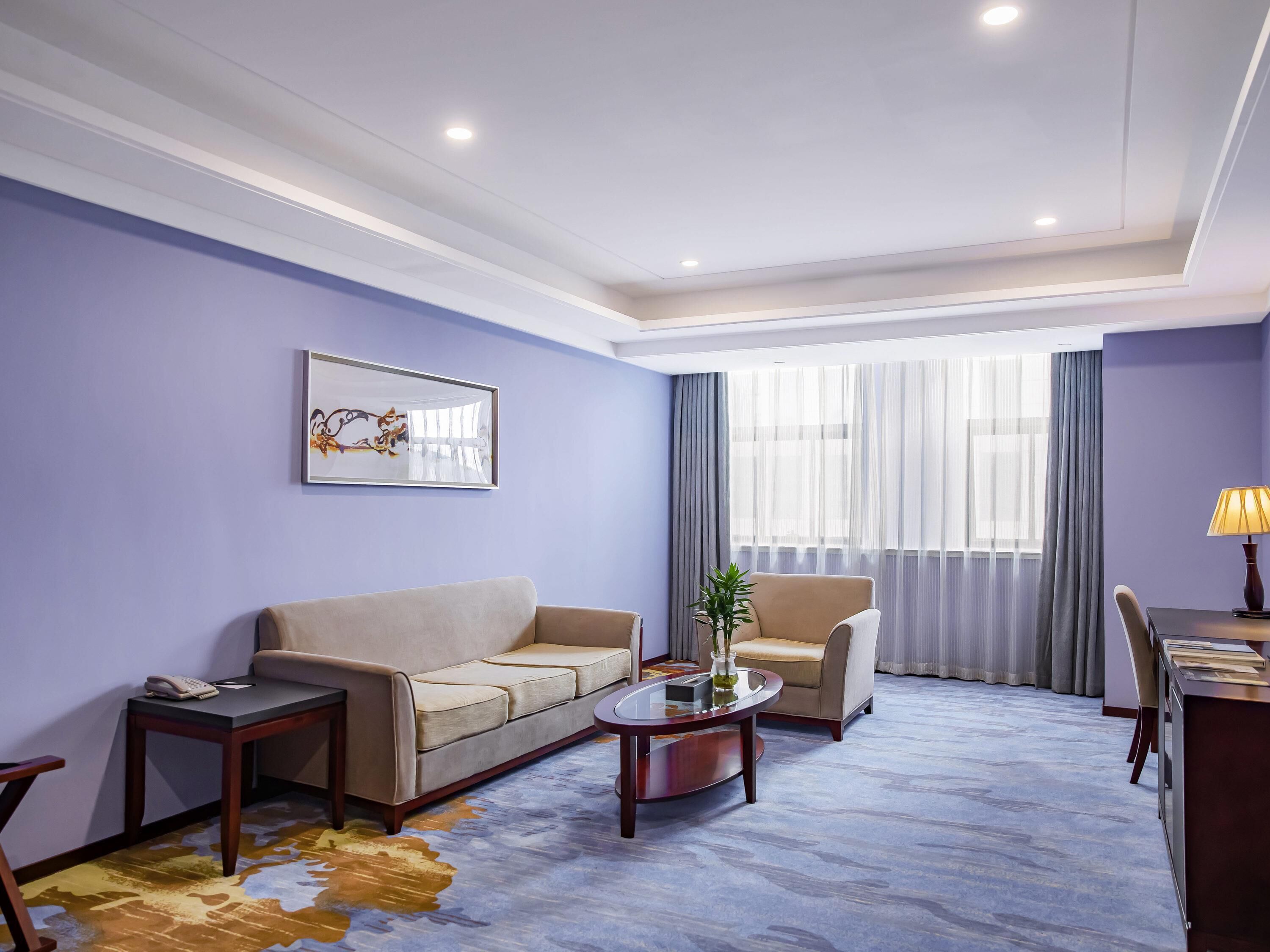Habitación de hotel con sofá, sillón, mesa de centro, escritorio, lámpara y un gran ventanal con cortinas.
