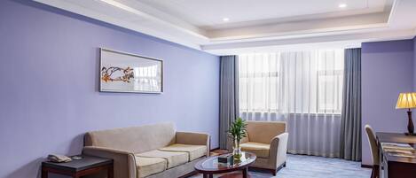 Habitación de hotel con sofá, sillón, mesa de centro, escritorio, lámpara y un gran ventanal con cortinas.