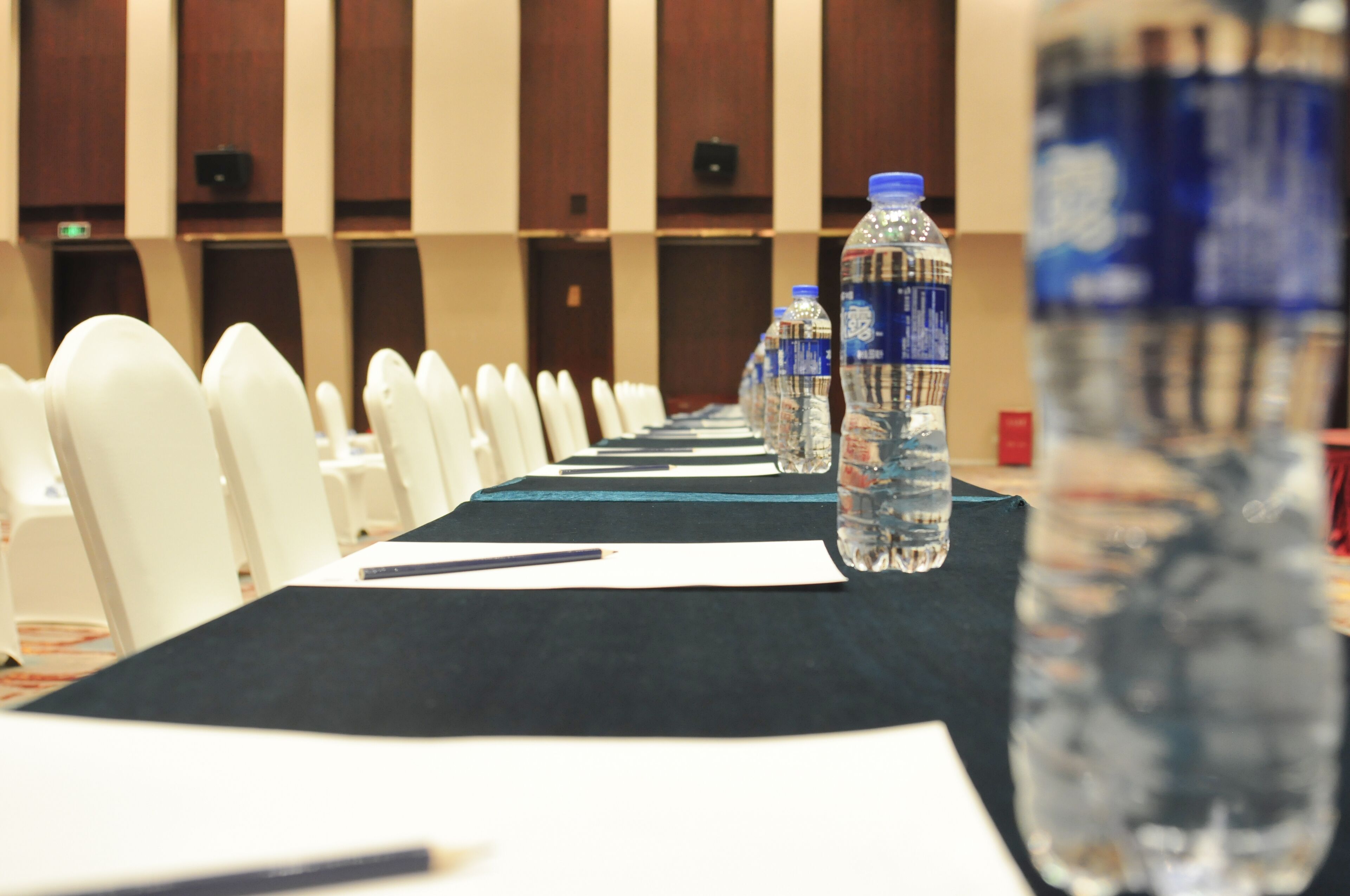 Un salón de conferencias con una larga mesa, sillas blancas, botellas de agua y un dispensador grande.