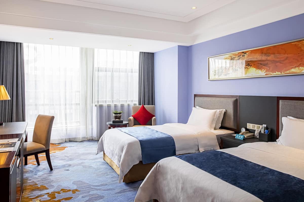 Ein Hotelzimmer mit zwei Betten, einem Schreibtisch, einem Stuhl und einem großen Fenster mit Vorhängen.