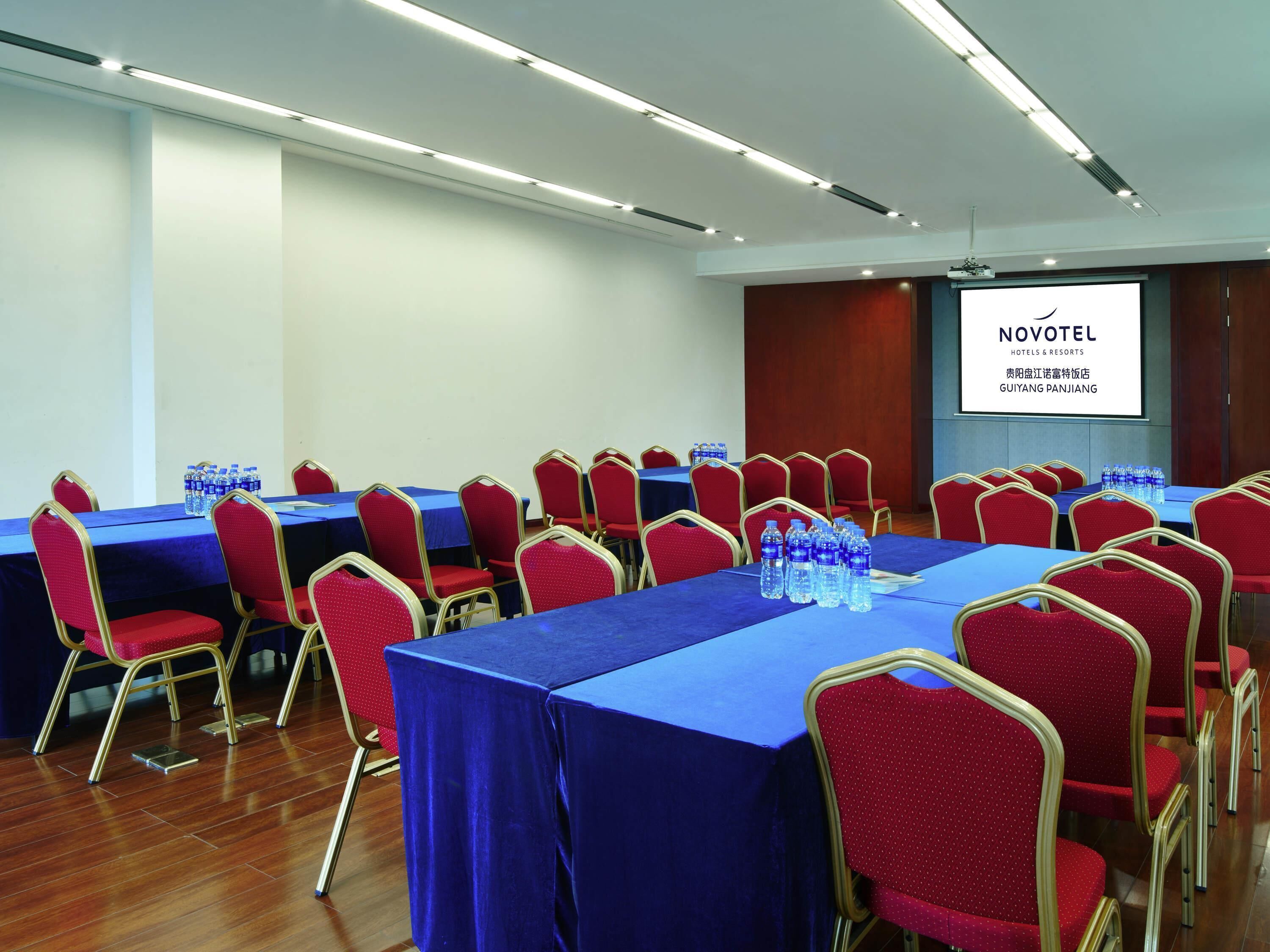 Un salón de conferencias con mesas y sillas, botellas de agua y una pantalla proyectando información del hotel.