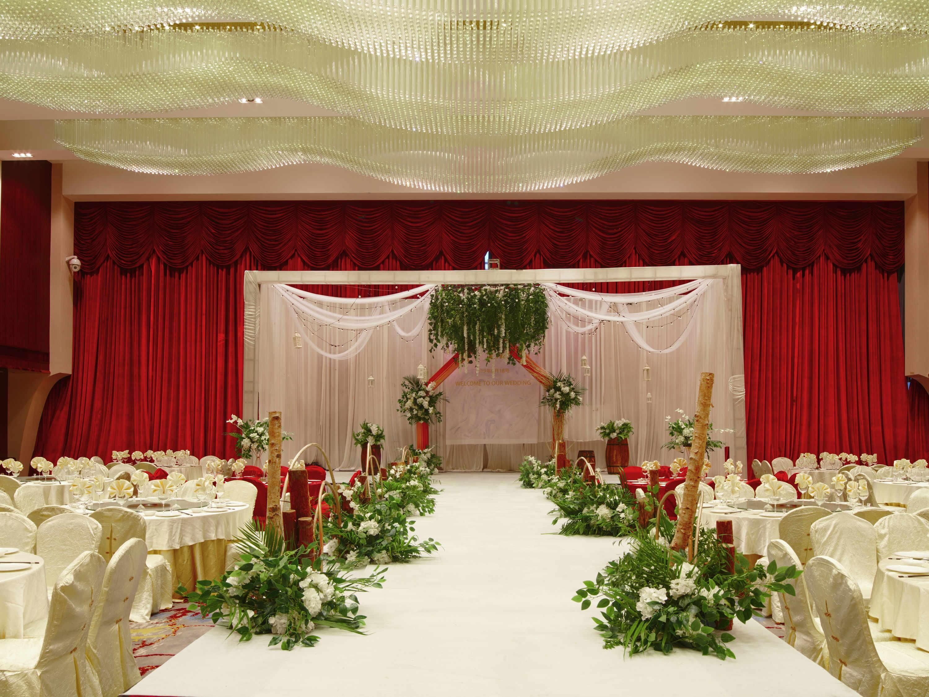 A banquet hall with rows of white tables and chairs, a stage with red curtains, and floral decorations.