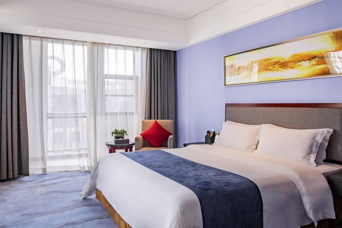 Ein Hotelzimmer mit einem großen Bett, einem Nachttisch, einem Sessel und einem Fenster mit Vorhängen.
